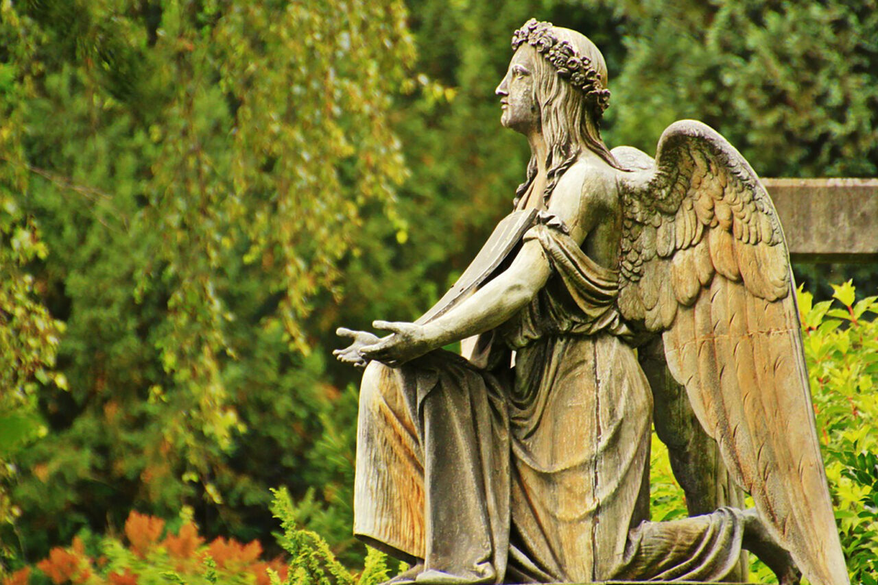 Ruskeansävyinen puusta veistetty polvillaan oleva enkelipatsas taustallaan risti.