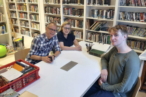 Kolme hymyilevää henkilöä istuu valkoisen pöydän ääressä taustallaan seinät peittävä kirjahylly.