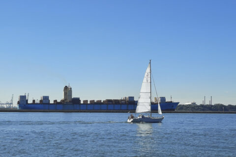 Sininen taivas ja meri, etualla purjevene ja taustalla rahtilaiva.