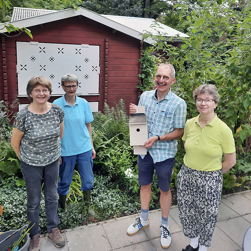 Kolme naista ja yksi mies, jolla on linnunpönttö käsissään, seisoo kesäisessä puutarhassa taustallaan punainen puurakennus.