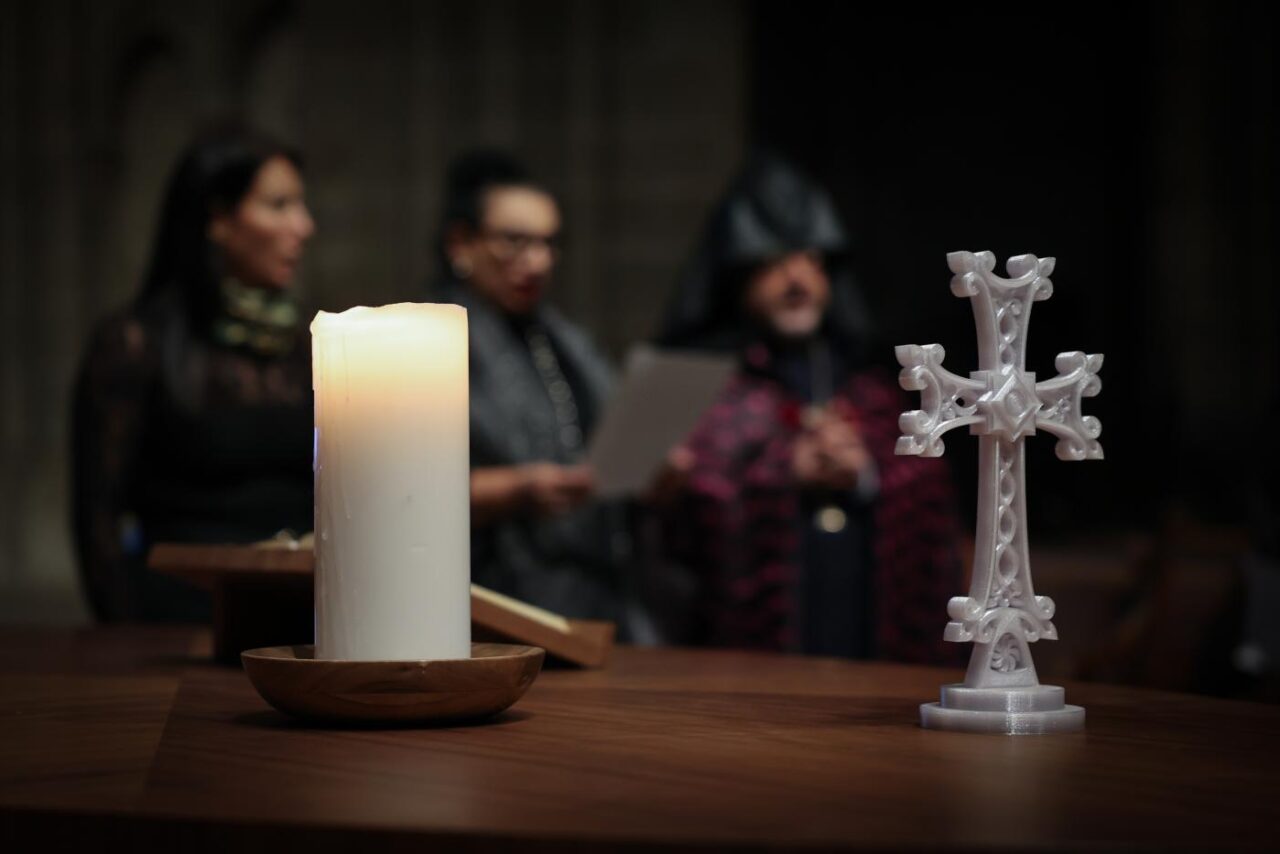 Kynttilä ja risti pöydällä. Taustalla kolme ihmistä istumassa.