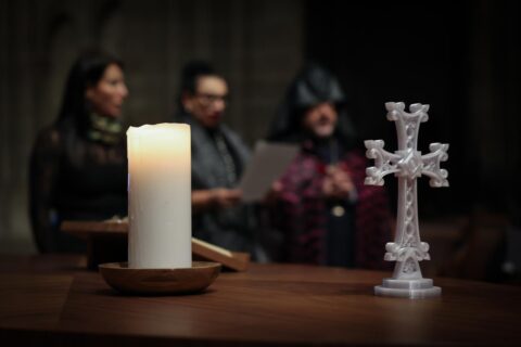 Kynttilä ja risti pöydällä. Taustalla kolme ihmistä istumassa.